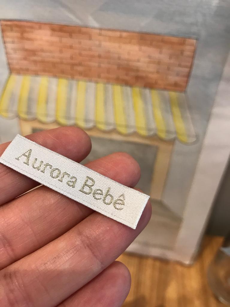 Etiqueta bordada Aurora Bebê nas mãos, diante de uma ilustração de loja.